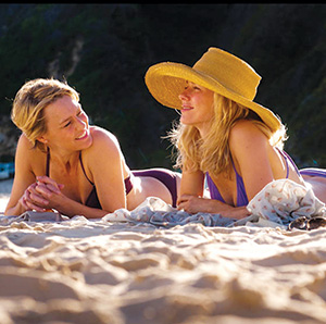 Two women in swimsuits lying on a sandy beach, smiling and talking to each other. One wears a large sunhat, and sunlight shines on both of them, with a blurred natural background.
