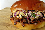 Close-up of a pulled pork sandwich on a wooden surface, featuring shredded pork, coleslaw with purple and green cabbage, and a glossy, golden-brown bun.