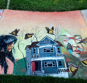 Colorful sidewalk chalk art depicts a blue house, a woman with long dark hair, a masked face, leafless trees, and several large butterflies on a pink and yellow background bordered by grass.