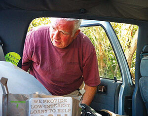 An older man with gray hair, wearing a maroon shirt, leans into the back of a car filled with bags and groceries. One bag reads, “We provide low-interest loans to help local producers.”.