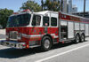 A red and white fire truck is parked on a street in daylight, with trees and buildings visible in the background. The truck has emergency lights on top and compartments on the side.