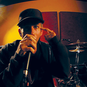 A person in a dark hoodie and cap holds a microphone, gesturing with one hand near their head, while standing in front of a drum set in a dimly lit music studio.
