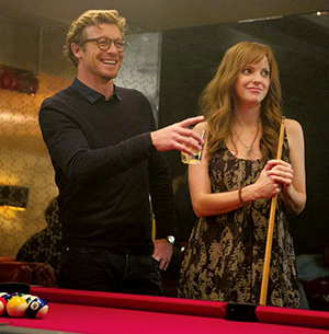 A man holding a drink and a woman holding a pool cue stand by a red pool table, both smiling and appearing relaxed in a dimly lit room.
