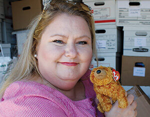 A woman with blond hair wearing a pink top smiles while holding a small, brown stuffed dog. She stands in front of shelves filled with boxes.