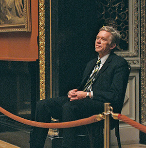 A man in a suit sits on a chair, appearing relaxed, inside an ornate room with framed artwork and gold accents visible in the background. A red rope barrier is in front of him.