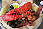 A seafood platter featuring a whole cooked lobster, corn on the cob, red potatoes, sausage, clams, and mussels, served on brown paper with cups of melted butter.