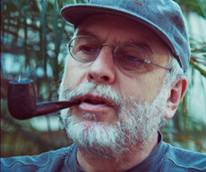 An older man with a gray beard and glasses wears a cap and holds a smoking pipe in his mouth. He is outdoors, with blurred greenery in the background.
