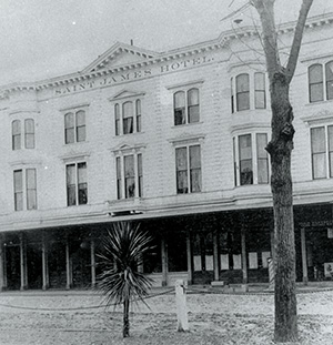 Historic black-and-white photo of the Saint James Hotel, a large white building with tall windows and a covered porch. A tree and a short, spiky plant stand in the foreground.