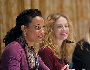 Two women sit at a table speaking into microphones during what appears to be a panel discussion. One woman in front gestures with her hands while the other woman next to her smiles.