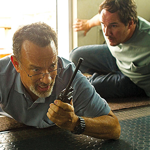 Two men are lying on the floor indoors. The man in front, wearing glasses, holds a walkie-talkie and looks tense. The man behind him appears alert, leaning on one hand and looking in the same direction.