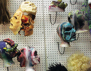 Several colorful, abstract masks and wigs are displayed on mannequin heads attached to a pegboard wall, with some wigs hanging below. The masks feature various textures and shapes.