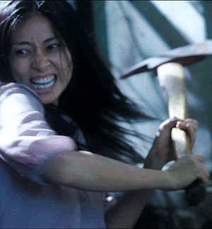 A woman with long dark hair swings a pickaxe with a fierce, determined expression, wearing a light-colored shirt in a dimly lit setting.