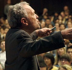 A man in a suit gestures with his hands while speaking to a large audience seated in an auditorium. The audience appears engaged and attentive.