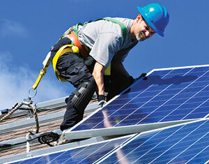 A worker wearing a blue hard hat and safety harness installs solar panels on a sloped rooftop under a clear blue sky.