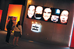 Two people stand in front of a wall displaying six illuminated, close-up portraits of faces with various expressions in an art gallery setting.