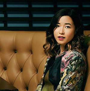 A woman with wavy dark hair and a colorful patterned jacket sits on a tan tufted booth, looking toward the camera with a slight smile. The background features dark horizontal slats.