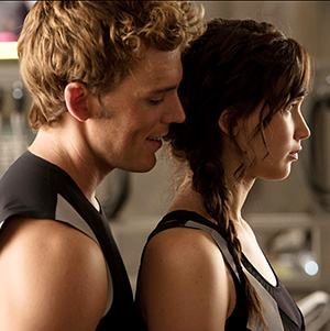 A young man with curly blond hair stands close behind a young woman with a dark braid, both wearing black and gray athletic outfits, in a softly lit indoor setting. The woman looks serious while the man smiles gently.