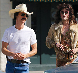 Two people stand outdoors. One wears a cowboy hat, sunglasses, a white T-shirt, and jeans. The other wears a fur coat, sunglasses, and holds a purse, with wavy hair and bracelets, standing beside a car.
