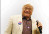 An older man with white hair and glasses smiles while holding a microphone. He wears a beige blazer, a striped shirt, and campaign buttons on his jacket. The background is plain and light-colored.