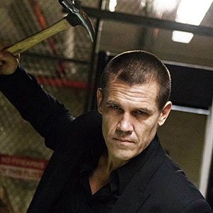 A man in a black suit with a serious expression raises a hammer above his head in a threatening manner, standing in an industrial or warehouse-like setting with metal fencing in the background.