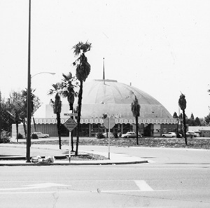 Black and white photo of a round, dome-shaped building surrounded by palm trees, with cars parked nearby and street signs visible in the foreground.
