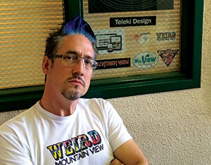 A man with glasses and blue-tinted hair stands with arms crossed, wearing a WEIRD MOUNTAIN VIEW t-shirt, in front of a window with signs for Teleki Design and other graphics.