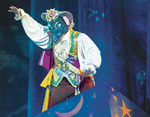 A performer in an ornate costume and a wolf mask stands atop a blue stage prop decorated with yellow stars, a crescent moon, and other celestial symbols, under dramatic stage lighting.