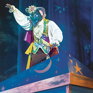 A performer in an ornate costume and a wolf mask stands atop a blue stage prop decorated with yellow stars, a crescent moon, and other celestial symbols, under dramatic stage lighting.