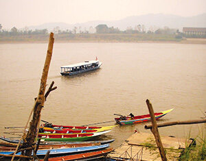 Wooden stairs lead down to a riverbank where several colorful longboats are docked. A larger boat floats on the river under a hazy sky, with distant hills in the background.