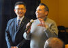 Two men stand indoors; one in a suit smiles while the other, wearing a gray sweater, speaks into a microphone and gestures with his hand.