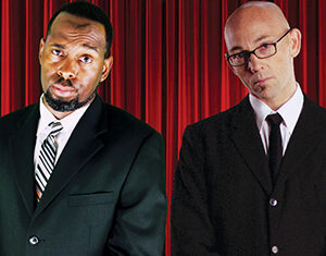 Two men wearing dark suits stand in front of red curtains. One has a beard and wears a striped tie, while the other is bald, wears glasses, and has a plain tie. Both have serious expressions and their hands clasped.
