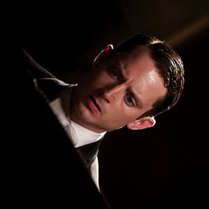A man with neatly styled dark hair, wearing a suit and tie, looks intently downward with a serious and focused expression in dim lighting.
