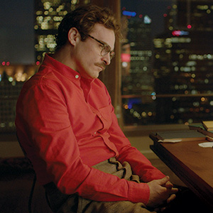 A man with glasses and a mustache, wearing a bright red shirt, sits pensively at a desk by a window overlooking a cityscape at night with blurred lights in the background.