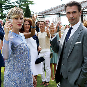 A well-dressed man and woman hold up champagne glasses at an outdoor gathering, with several smiling people in the background also raising glasses, celebrating on a sunny day.