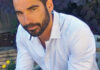 A man with dark hair and a beard, wearing a light blue button-up shirt and blue pants, sits outdoors on a bench near flowers, looking at the camera with a slight smile.