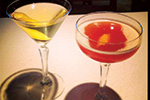 Two cocktails in stemmed glasses on a light surface; one drink is pale yellow with a lemon twist, the other is reddish-orange with an orange peel garnish.