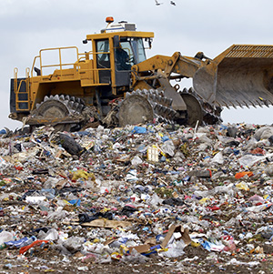 A large yellow bulldozer moves and compacts piles of mixed trash and debris at a landfill under a cloudy sky.
