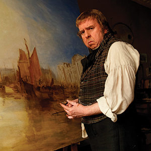 A man in historical clothing stands in front of a large painting of boats on water, holding painting tools, with a serious expression on his face.