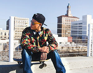A man wearing a colorful shirt, jeans, black sneakers, headphones, and a backward cap sits on a skateboard in an urban setting with buildings and a clear sky in the background.