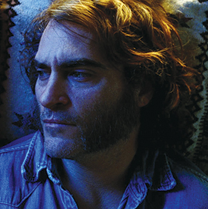 A man with wavy hair and sideburns lies on his back, looking to the side. His face is partially illuminated with blue light, and he wears a denim shirt. The background shows a patterned fabric.