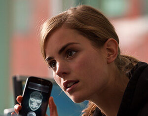 A young woman with light brown hair holds up a smartphone displaying a glowing face scan, looking intently at someone off-camera in a brightly lit indoor setting.