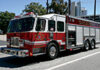 A red and white fire truck with emergency lights on top is parked on a street in an urban area, with buildings and trees in the background.