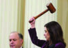 A woman in a purple jacket raises an oversized gavel, preparing to bring it down, while a man in a suit sits beside her with a serious expression. Curtains hang in the background.