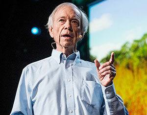 An older man in a light blue shirt speaks on stage, holding a remote and wearing a headset microphone, with a nature scene displayed on a screen behind him.