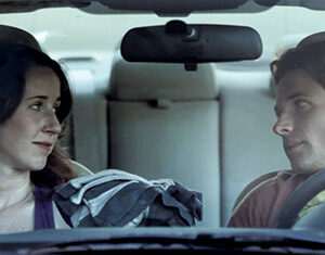 Two people sit in the front seats of a car, facing each other and sharing a serious, thoughtful look. The photo is taken from outside the car through the windshield.