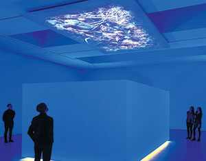 People stand in a dimly lit blue room surrounding a large, glowing, cube-shaped structure. Above the cube, abstract light patterns are projected on the ceiling, creating a futuristic and immersive atmosphere.