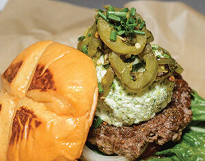 A hamburger with a brioche bun, leafy lettuce, onion, beef patty, a large dollop of herbed cheese, sliced jalapeños, and chopped chives, served on brown parchment paper.