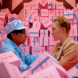 Two people face each other in front of a large stack of pink boxes with blue ribbons labeled Mendls. One wears a light blue bakery uniform and hat, the other has blonde hair styled in a braid.