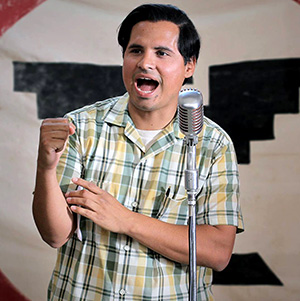 A man in a plaid shirt speaks passionately into a vintage microphone, gesturing with his hand. A large banner with bold, black designs is visible in the background.
