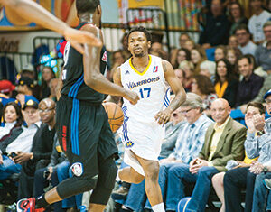 A basketball player in a white Warriors jersey, number 17, dribbles the ball down the court while a defender in a black jersey guards him. Spectators watch the game from the stands in the background.